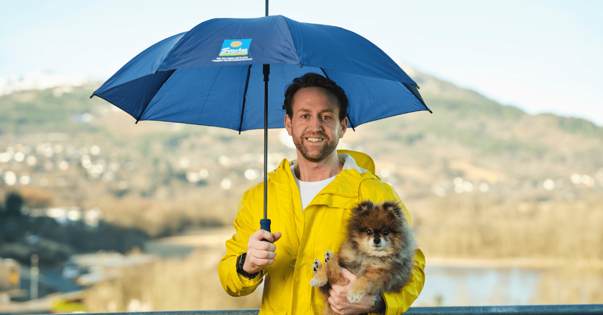 Bilde av mann med gul regnjakke og blå paraply med liten hund i armene. Begge ser mot kameraet og smiler. 