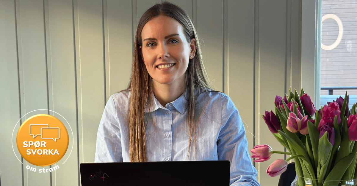 Bilde av kvinne som sitter og smiler mens hun ser mot kameraet. Hun har en laptop foran seg og man ser en bukett med tulipaner i høyre hjørne. 