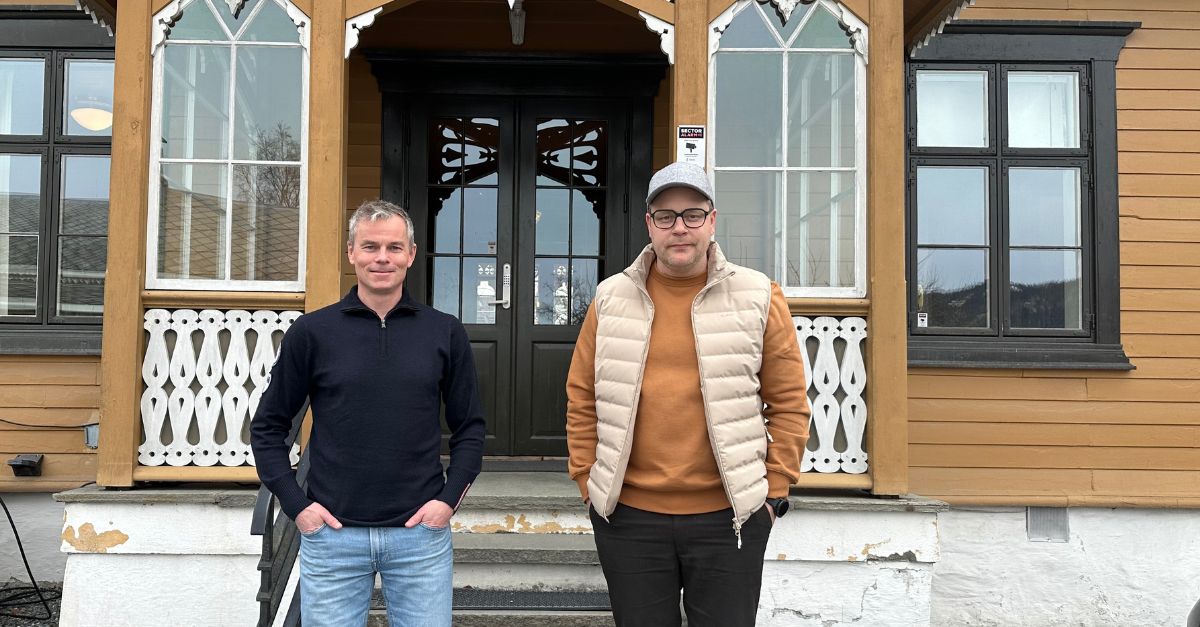 BIlde av to menn som ser mot kameraet foran trapp og inngangsparti på et gammelt og ærverdig hus. 