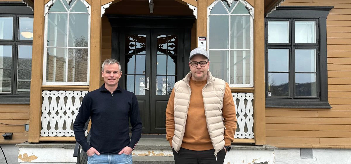 BIlde av to menn som ser mot kameraet foran trapp og inngangsparti på et gammelt og ærverdig hus.