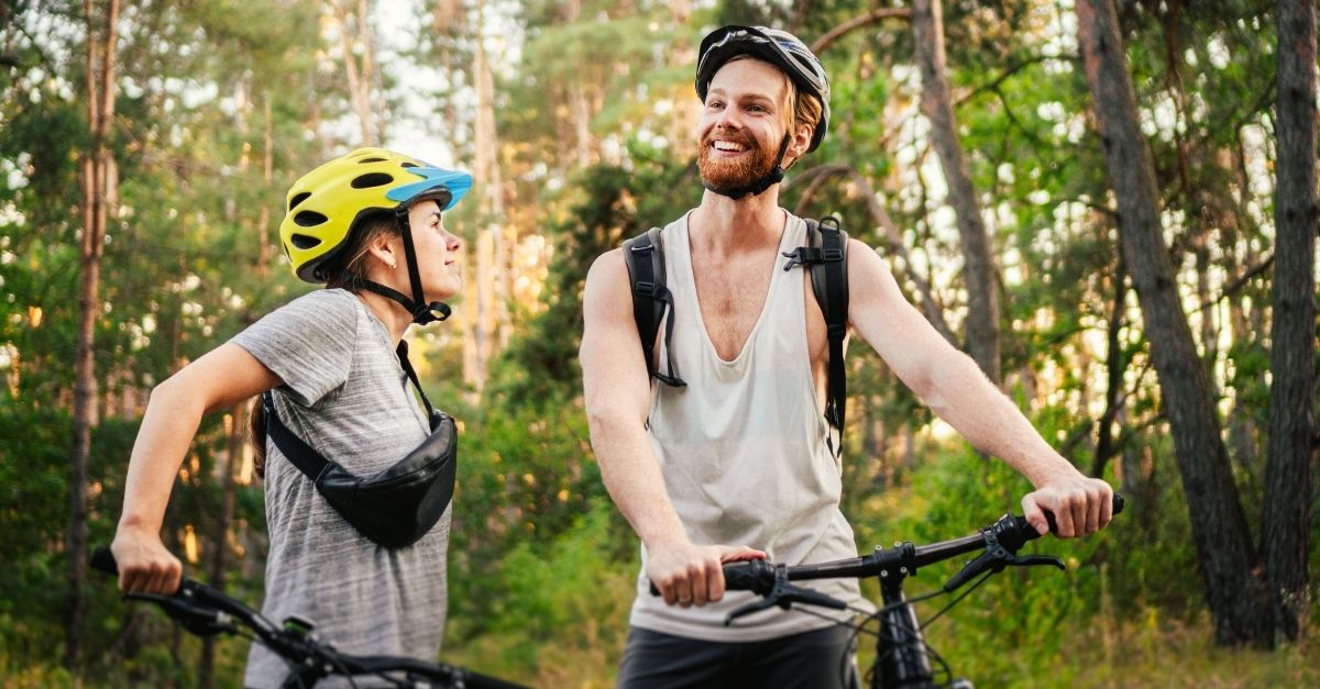 En smilende mann og kvinne står i skogen med hver sin sykkel og med hjelm på hodet.