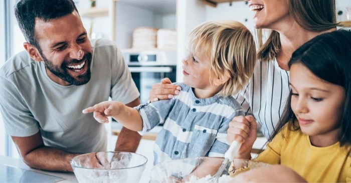 Familie på et kjøkken. Far smiler, mor og to barn ser på han. Boller med mel på benken foran dem.