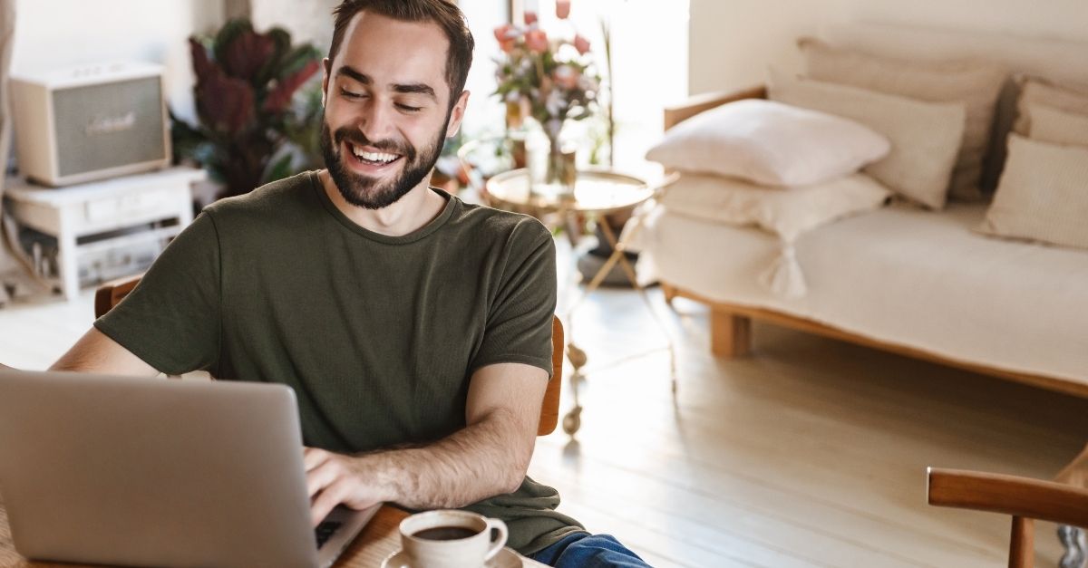 Mann sitter foran PC og smiler, han ser på skjermen. I bakgrunnen er det en lys sofa, lite sidebord med blomster og et musikkanlegg. 