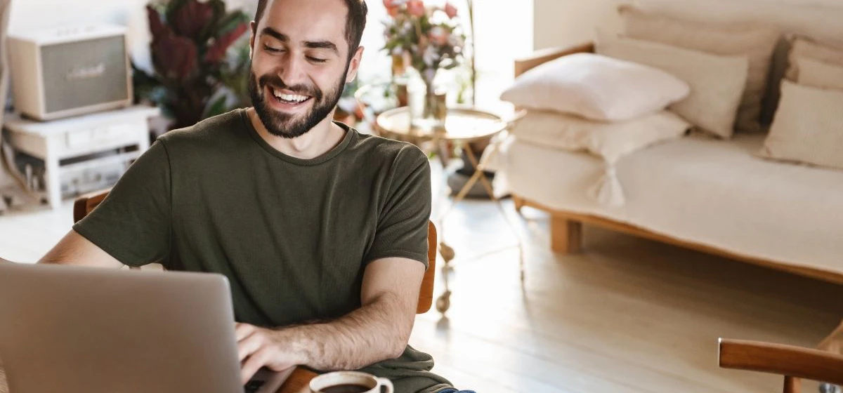 Mann sitter foran PC og smiler, han ser på skjermen. I bakgrunnen er det en lys sofa, lite sidebord med blomster og et musikkanlegg.