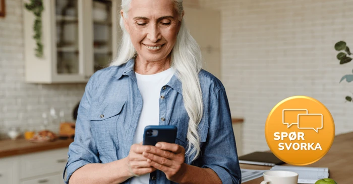 Kvinne står på kjøkkenet og ser på mobilen hun holder i hendene.